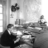 An undated photograph of Frederick C. Lincoln at his desk in Washington, with aluminum bird bands and pliers to his right and a map of North American migratory pathways on the wall. 